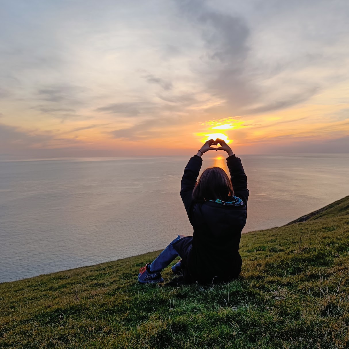 Person making a heart shape with hands against a Welsh coast sunset